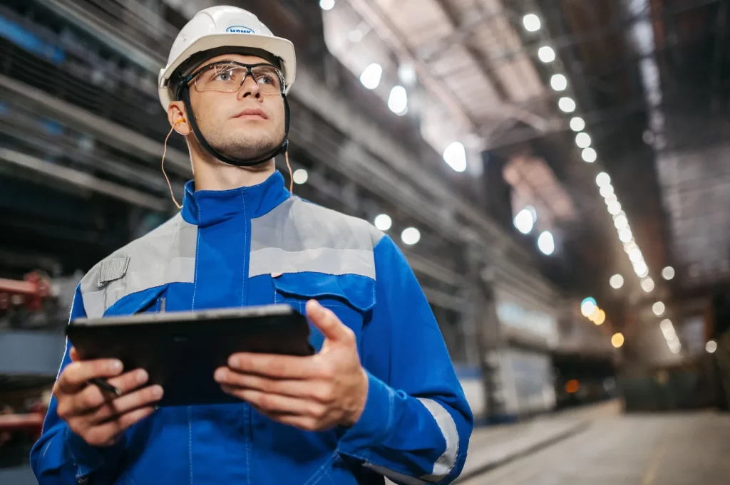 Pekerja pabrik berseragam safety memegang tablet di lantai produksi, ilustrasi aplikasi inspeksi kualitas untuk industri manufaktur yang menggantikan checklist kertas