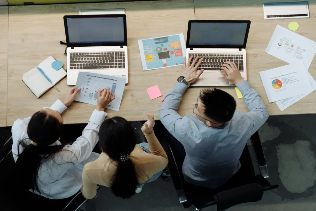 Tiga rekan kerja membahas laporan kinerja dan KPI di depan laptop, ilustrasi penggunaan aplikasi penilaian kinerja karyawan di perusahaan